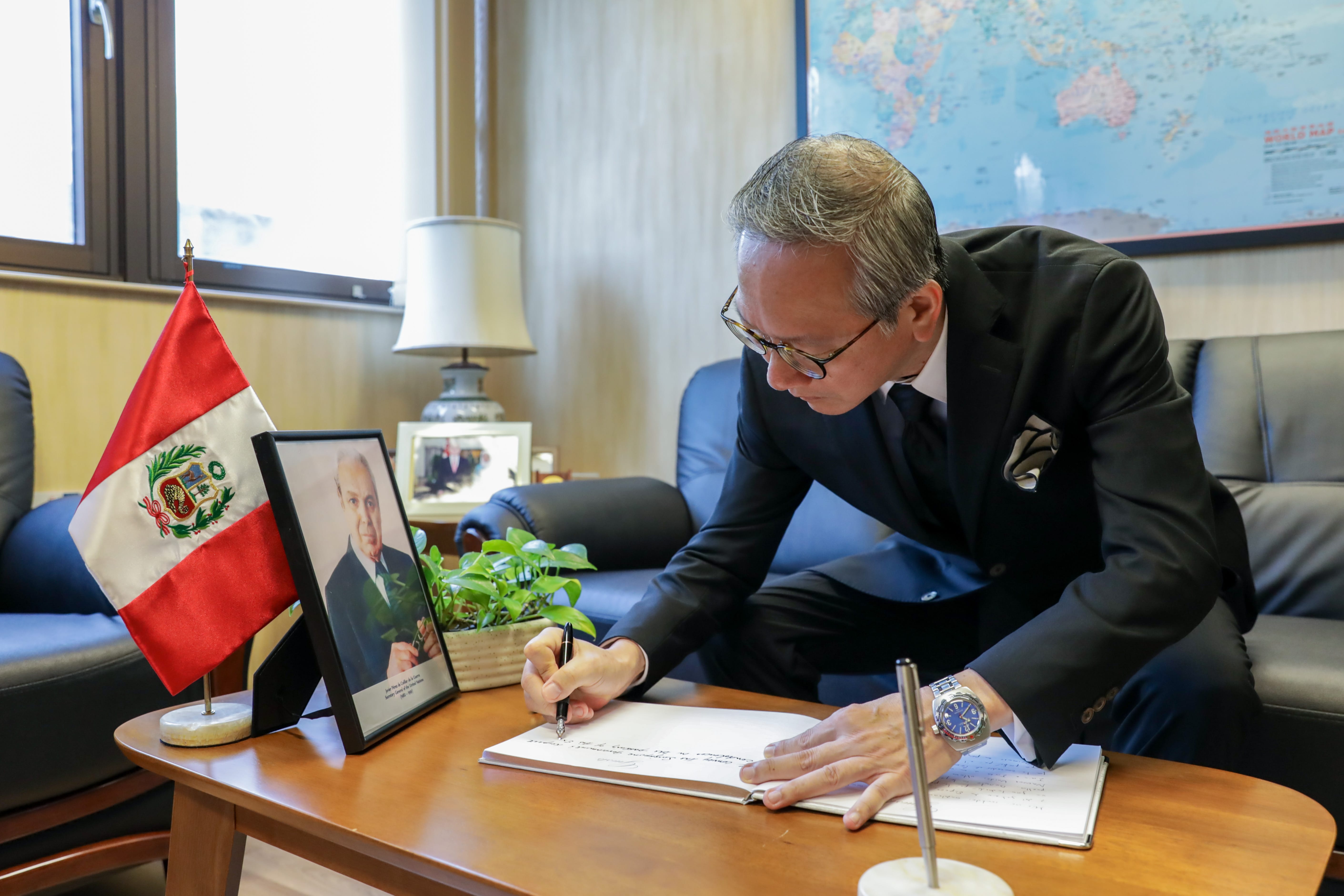 Man in suit signs document at desk with Peruvian flag and portrait of Javier Pérez de Cuéllar.